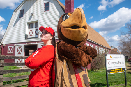 A young female in a red hat and red shirt stands back to back with the UMD mascot Testudo in front of the Campus Farm. 