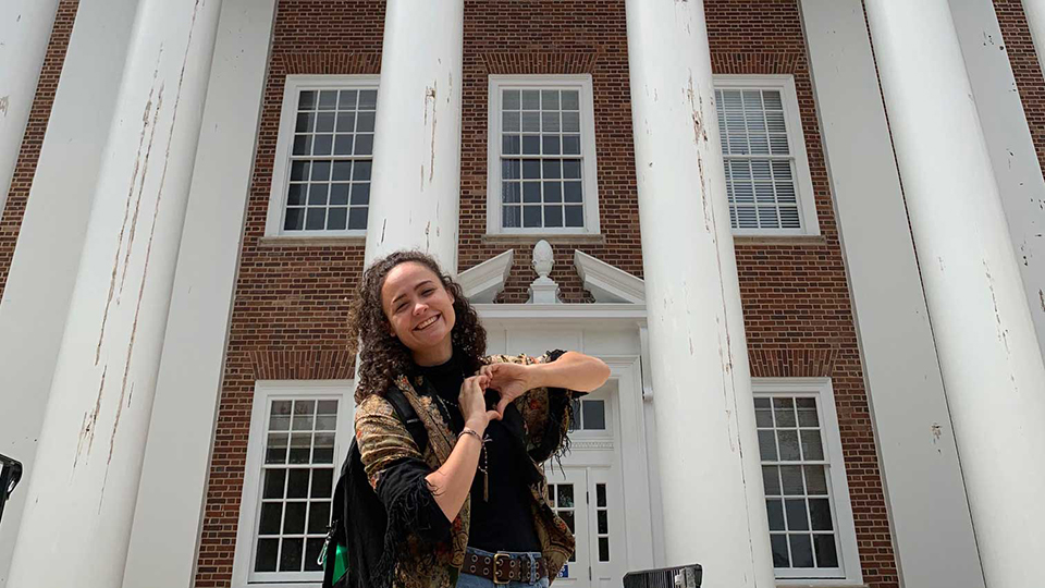 Female student standing in front of Symons Hall doing the heart symbol