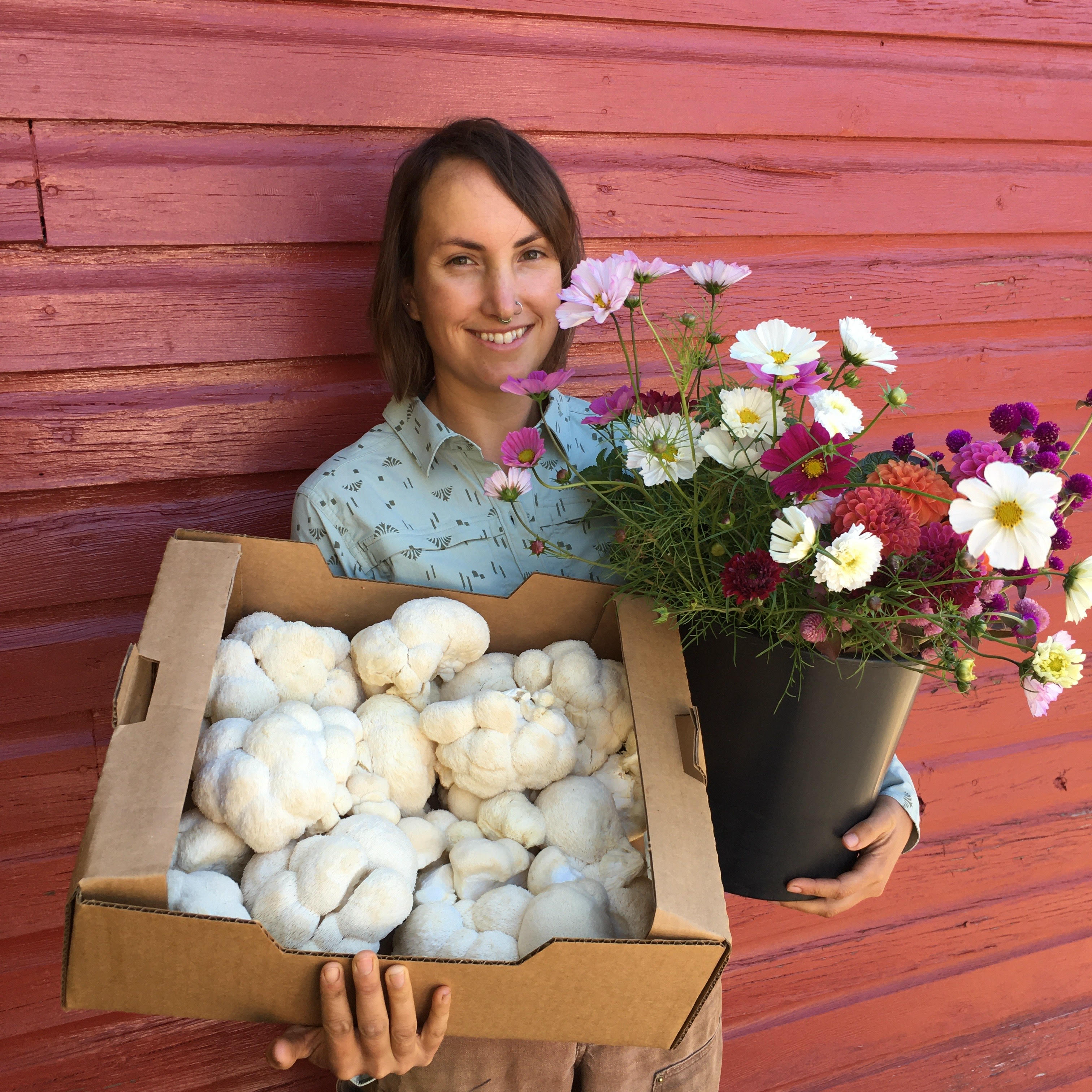 Farmer Courtney Cohen holding a box of mushrooms in her right hand and a put of flowers in her left hand.