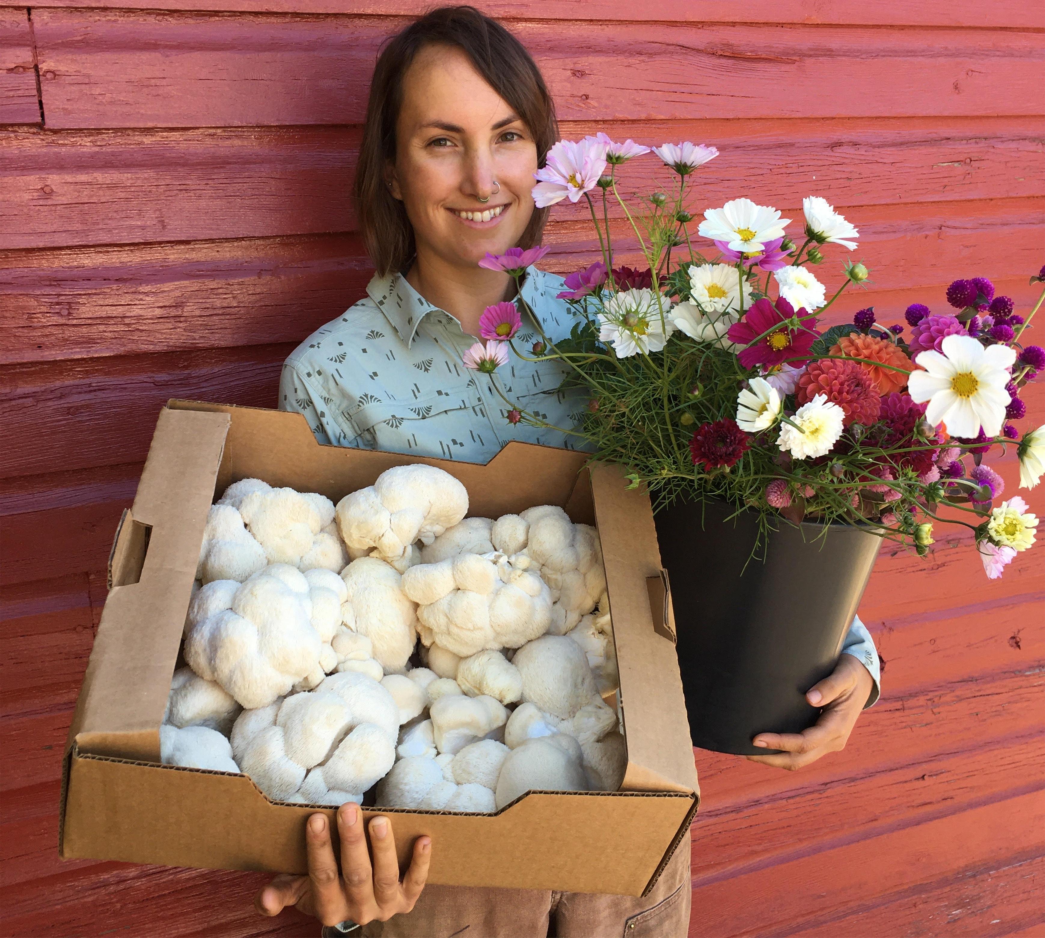 Farmer, Courtney Cohen, holding a box of mushrooms in her right hand and a pot of flowers in her left hand.