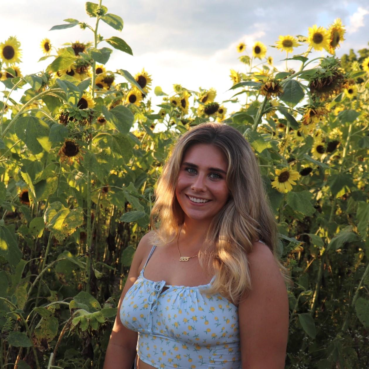 Grace is wearing a light blue shirt and standing in a field of sunflowers