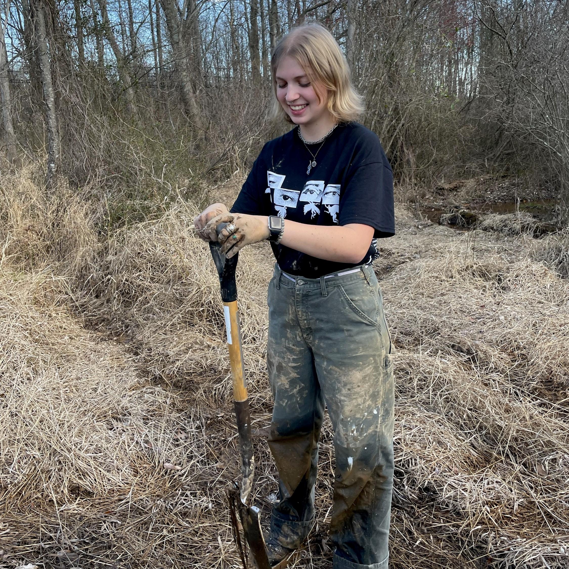 Caroline is holding a shovel and smiling while working with soil in a wetland