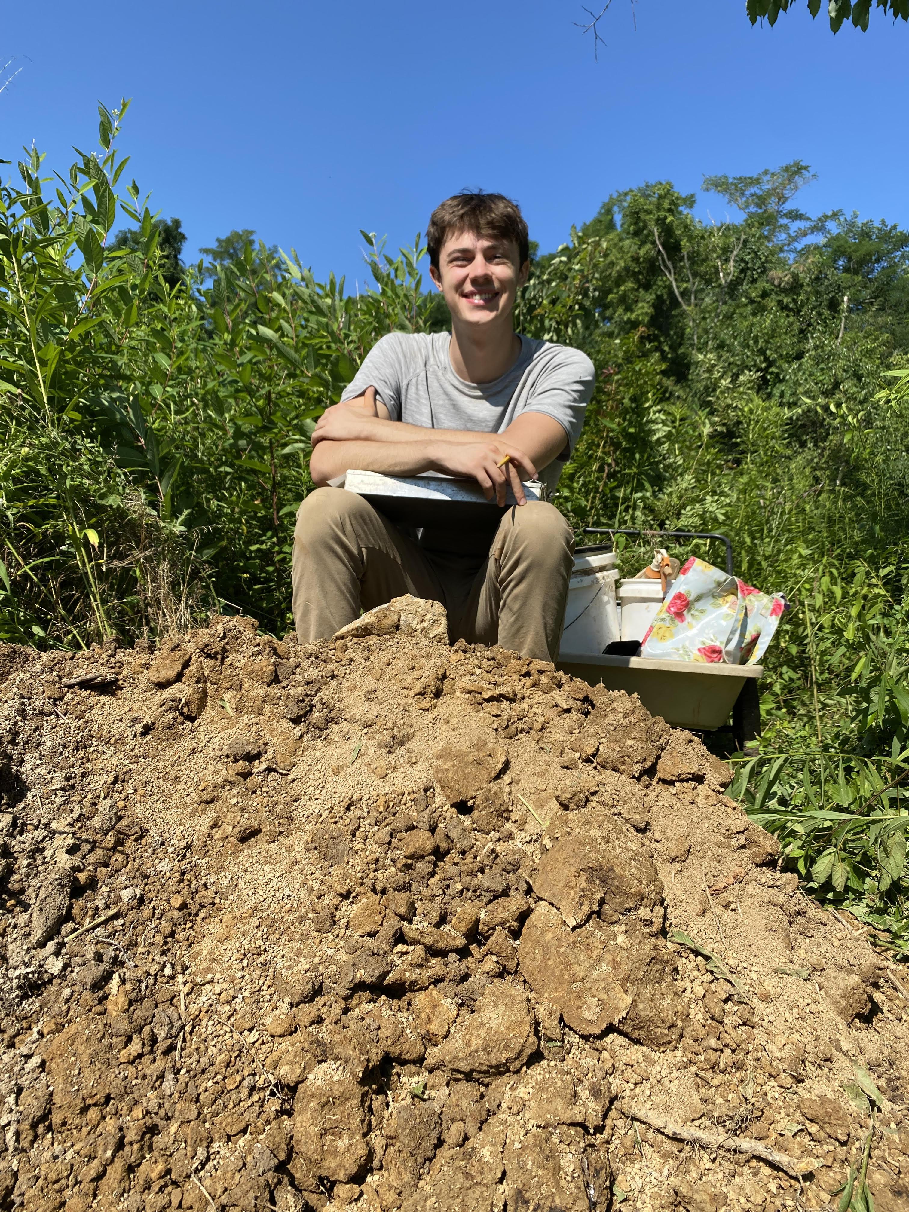 Alex Quigley. Happy student sitting above soil pit, accompanied by tools to help classify soil.