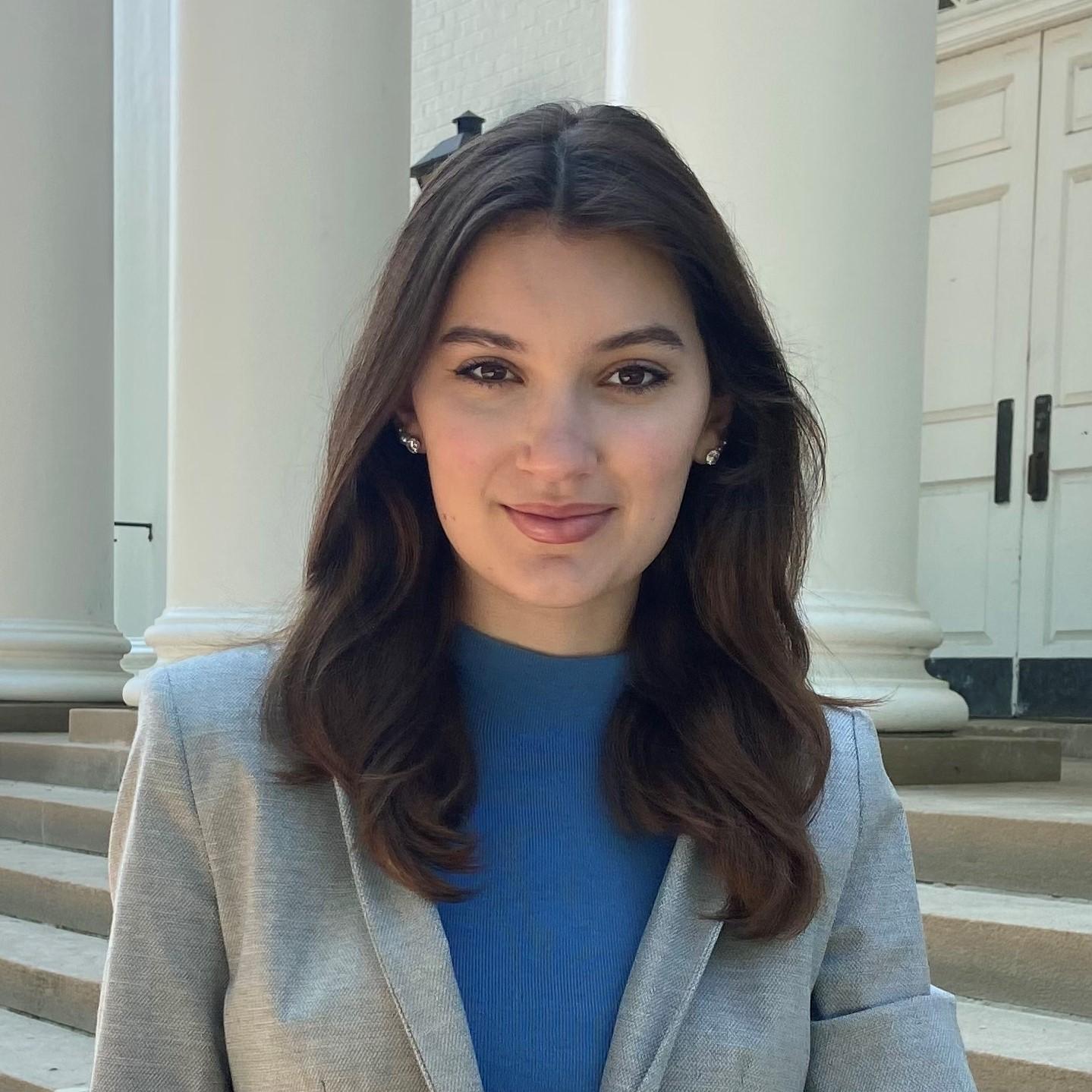 Maria is wearing a blue top and grey blazer, posing in front of pillars on campus