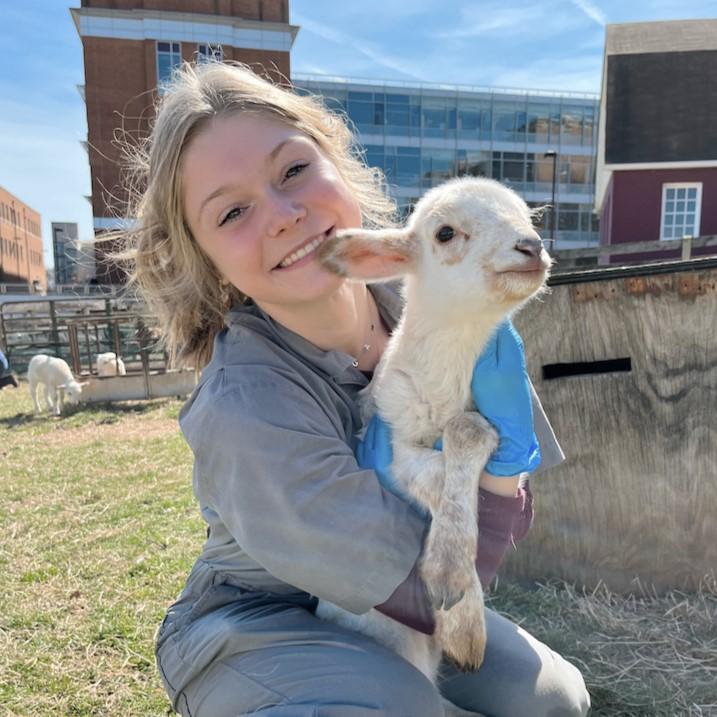 Megan is dressed in grey overalls, smiling and hugging a lamb on the campus farm
