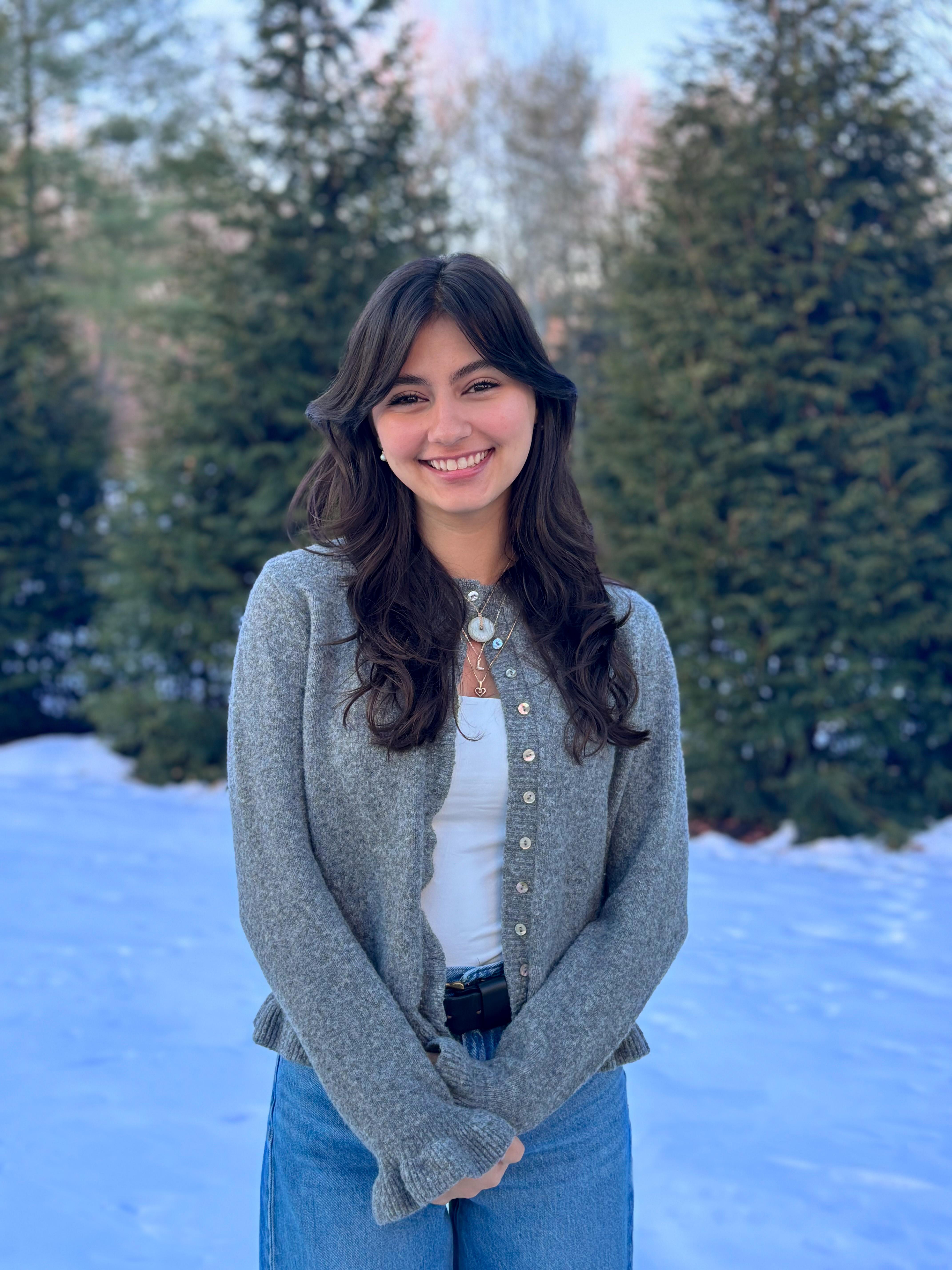 Friendly student in a sweater stands in the foreground of a snow-covered coniferous backdrop.