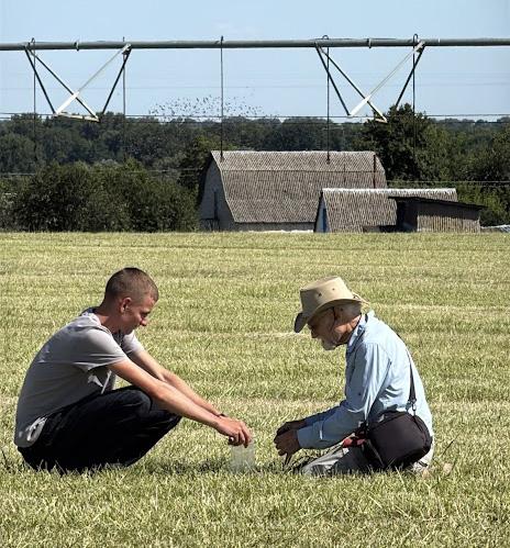Weil and a young man sit cross legged, facing each other in a field, working over something in Weil's hands. A large farm irrigator looms in the background.