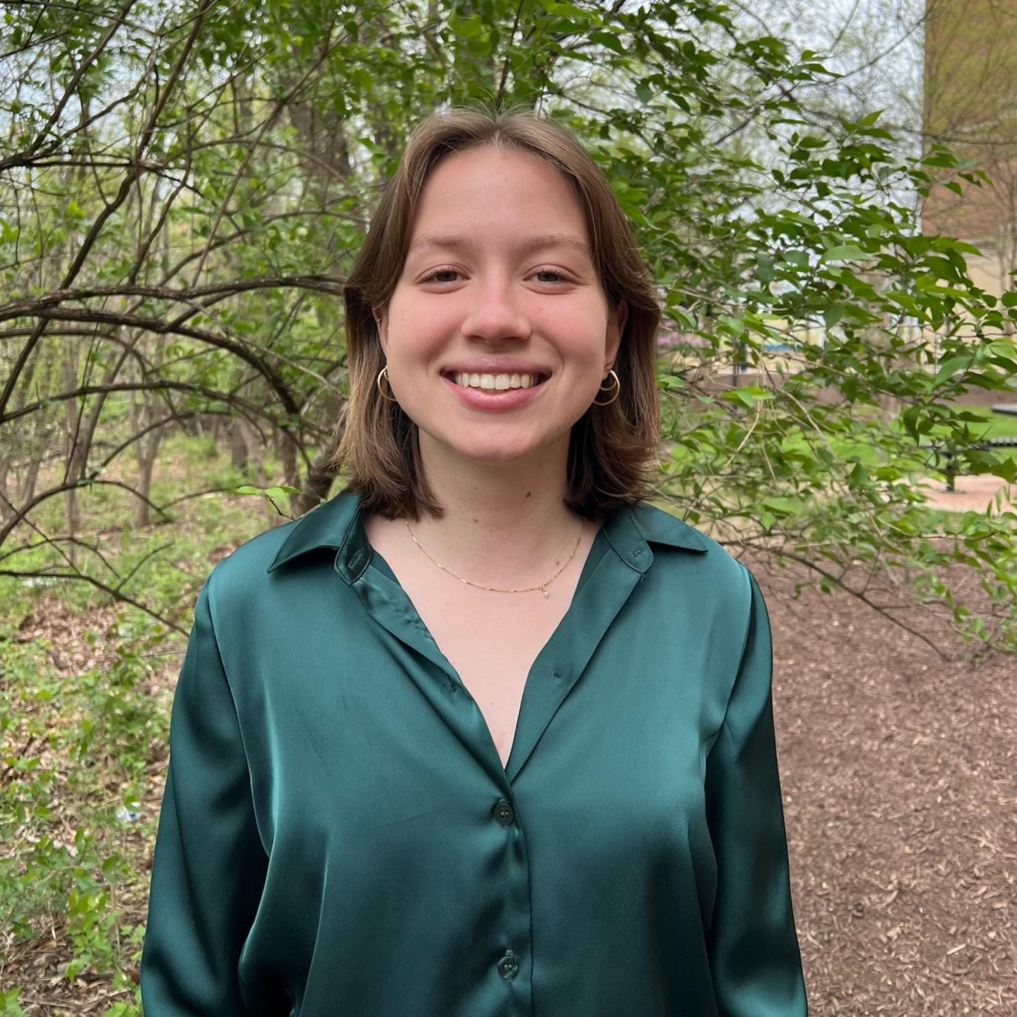 Adeline poses, smiling, in front of a tree. She is wearing a dark green blouse.