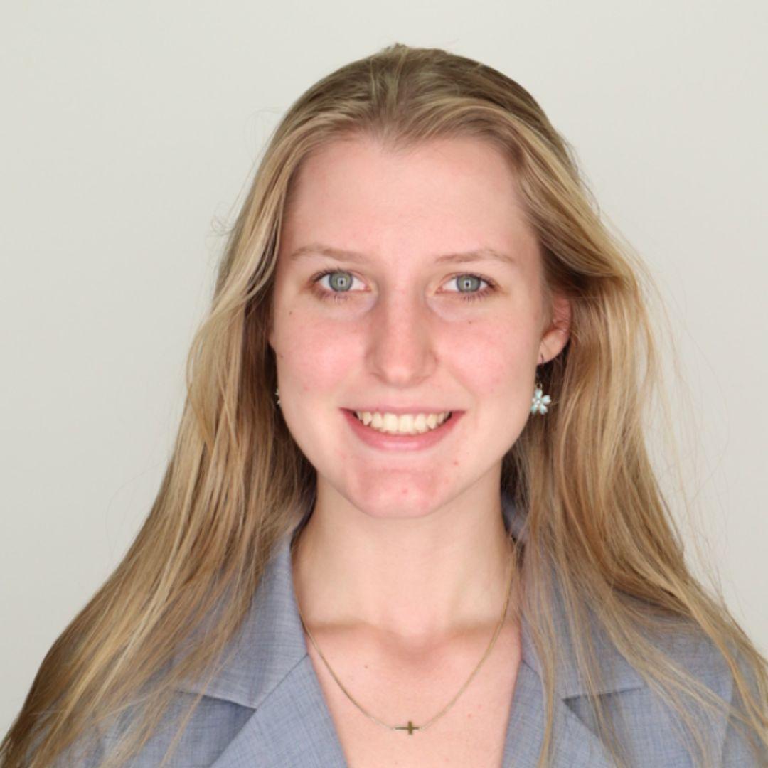 Izzy is wearing a grey blazer while smiling brightly at the camera