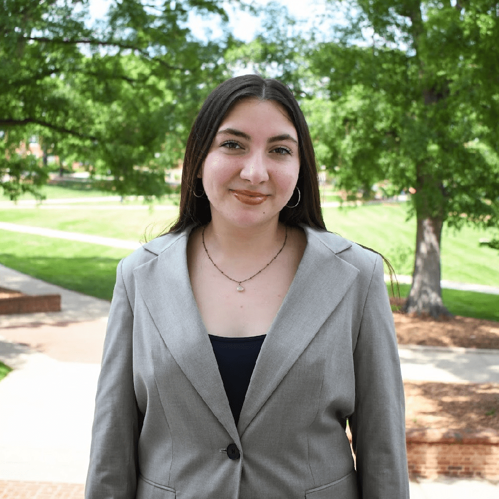 Maddie is wearing a black shirt and grey blazer, smiling at the camera near McKeldin Mall