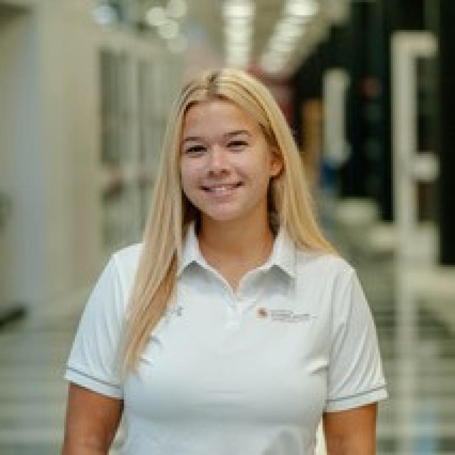 Cassidy is wearing a white polo and smiling for the camera with a long hallway in the background