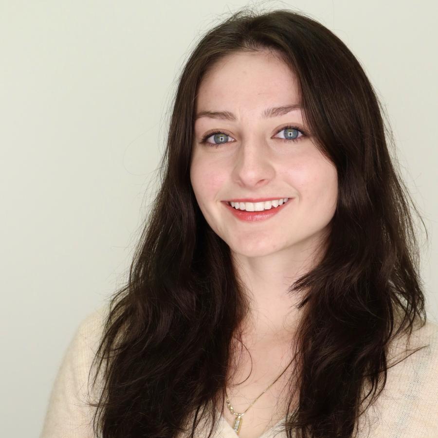 Katrina is wearing a beige sweater, smiling at the camera while standing in front of a white photo backdrop.