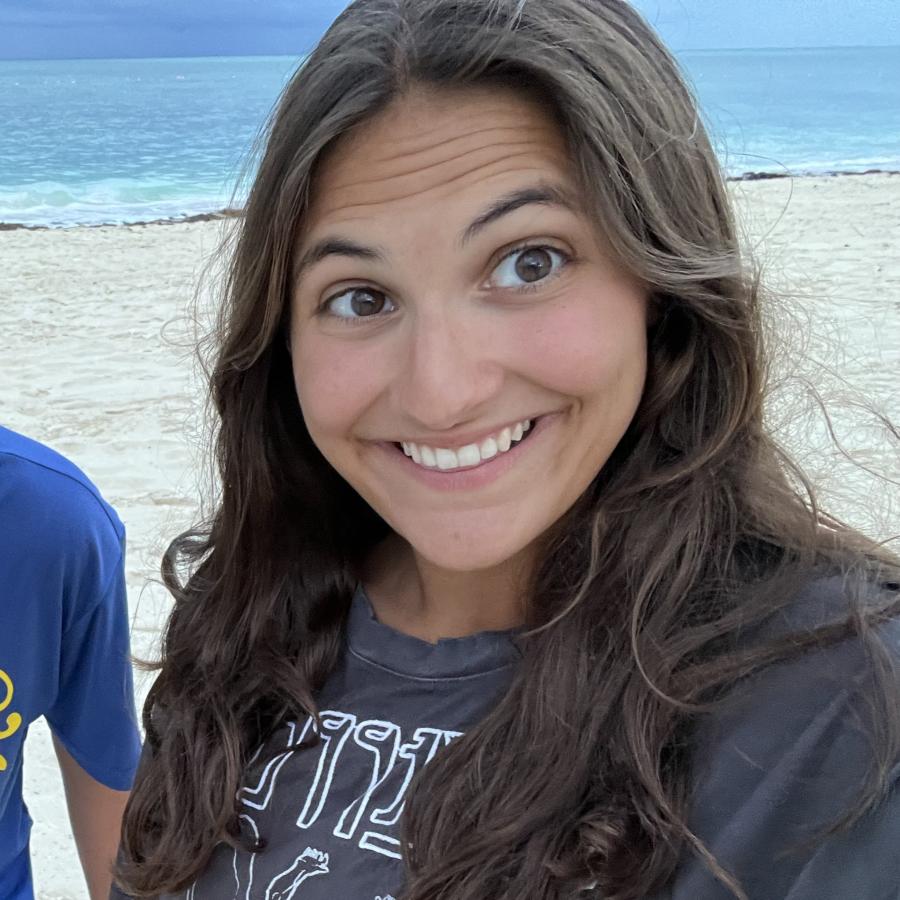 Cecilia is wearing a black tee shirt and smiling for the camera while standing on a beach with the ocean in the background.
