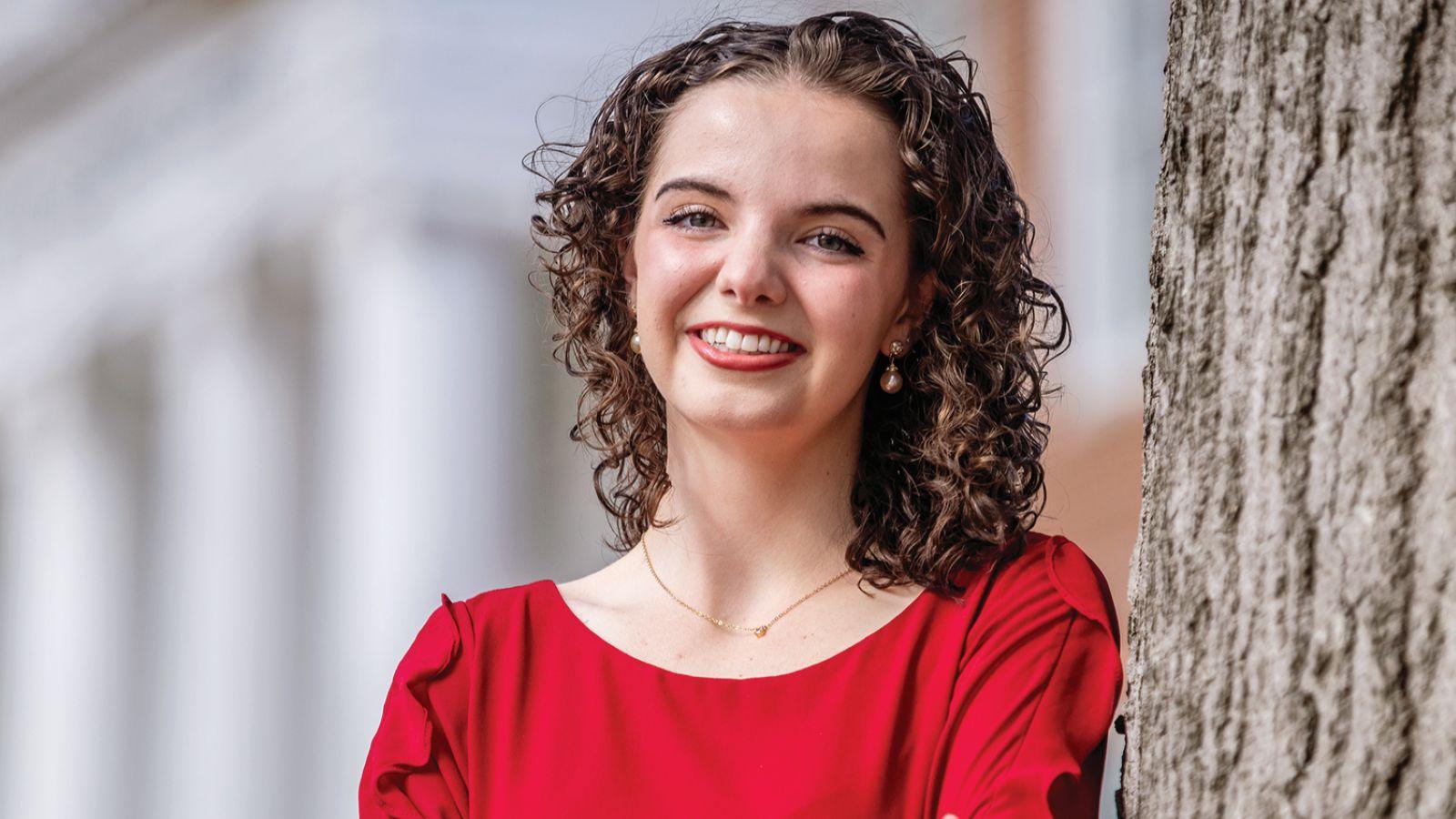 Esther Bonney wearing red smiling standing next to a building and a tree