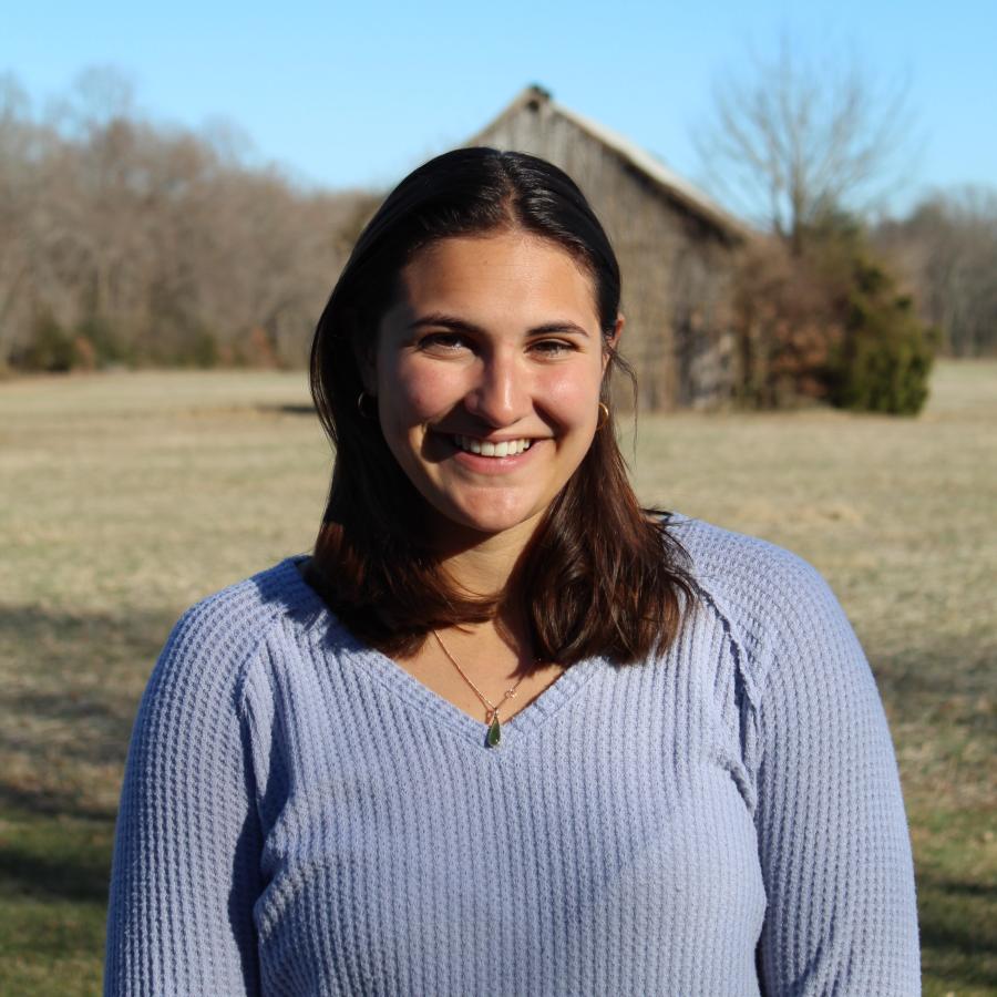 Cecilia is smiling and wearing a light blue sweater while standing in a field with an old barn in the background
