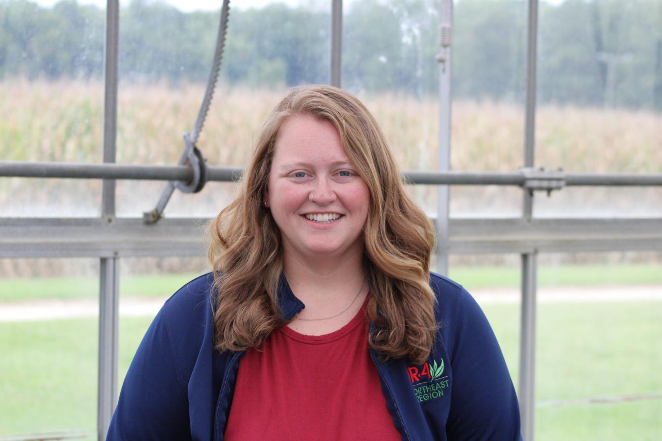 Woman named Megan stands in a greenhouse with a field of corn behind her.