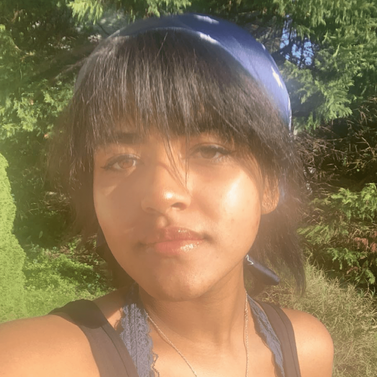 Kaity is standing in a wooded area, wearing a black tank top with a blue scarf in her hair